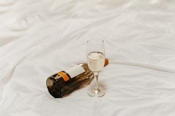 a bottle of wine and a beautiful glass goblet on the bed with pure white crumpled pastel linen in...
