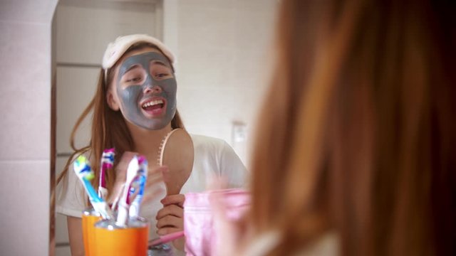 A young attractive woman with a mask on her face dancing and singing in front of the mirror in bathroom and having fun