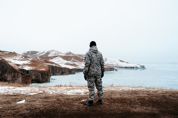 man in military clothing camouflage against the background of a winter landscape and pure white snow, a hunter stands back on amazing northern landscape, ice sea and hills in snow and ice