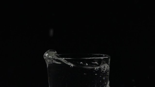 Slow motion close-up of ice cube falling down in a glass of water on black background with splash, bubbles and drops