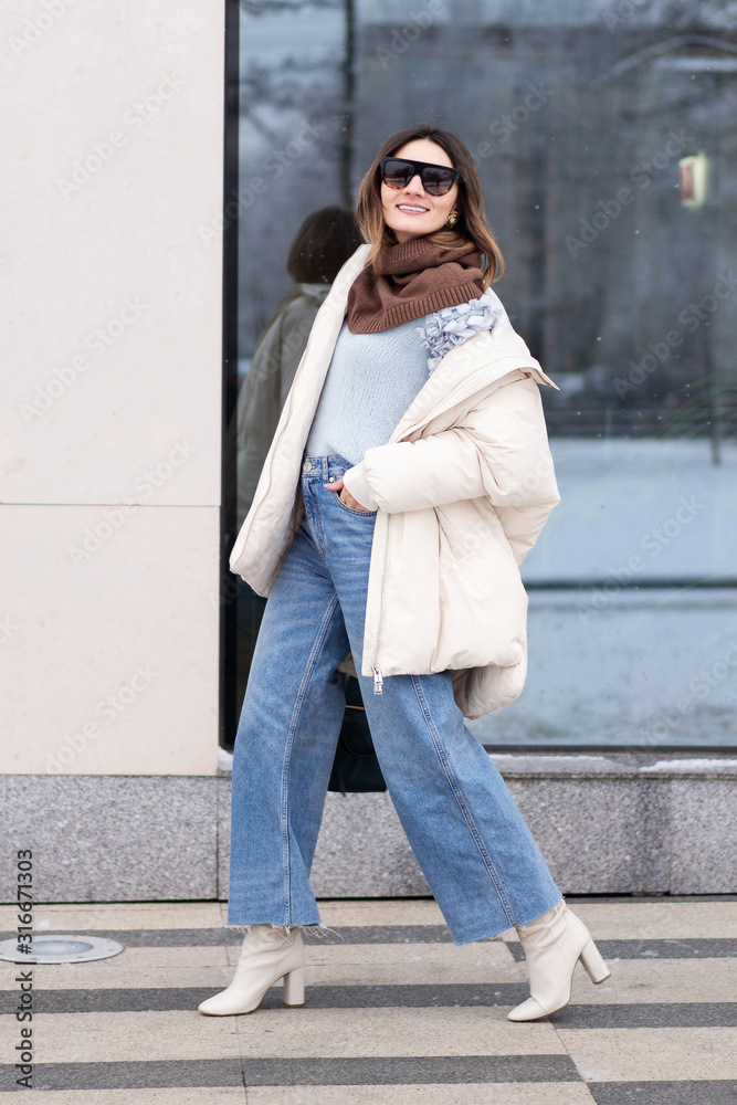 Wall mural European model girl in a beige oversized down jacket, knitted sweater, flared jeans with a handbag and glasses is walking down the street. Life style