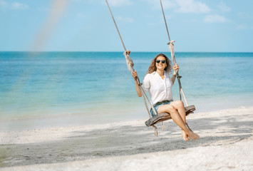 Beautiful young curly funny girl on a swing on a tropical beach, - obrazy, fototapety, plakaty