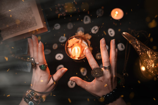 Astrology and esotericism. Female witch hands conjure over a candle. On a black background lie fortune-telling runes, a book, precious amulets, a copper lamp and a candle. Dark magic