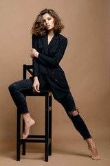 beauty fashion model with clean skin and curly hair in black jacket on biege background on the chair, serious business woman
