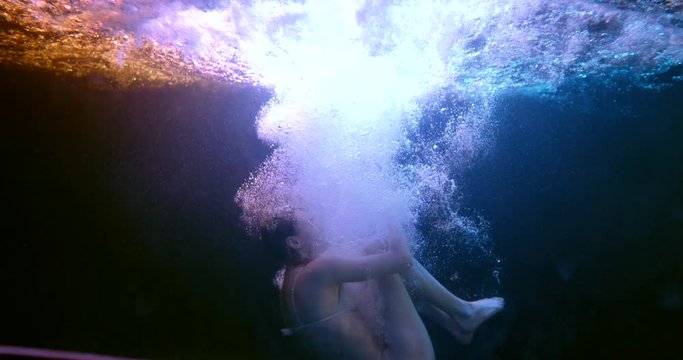 a girl in lingerie falls under the water in the fetal position in a stream of bubbles. then she floats up. the waves are on the surface. blue and orange backlight