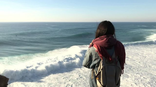 Young brunette girl standing on cliff edge, taking photo, enjoying a view of crushing waves of Nazare, Portugal; sunny windy evening, sunset time, amazing natural tourist attraction; travel concept