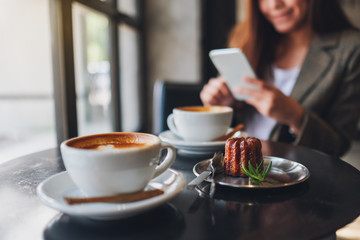 Closeup image of an asian woman holding and using mobile phone with coffee cups and snack on the table in cafe