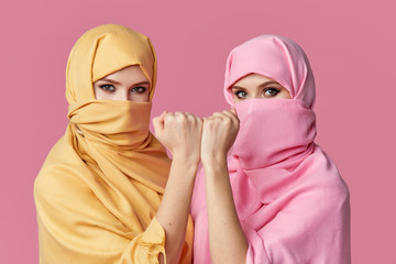 Girl power. Two young Muslim arabian women wearing colorful hijabs against pink background. Points finger to side. Space for text