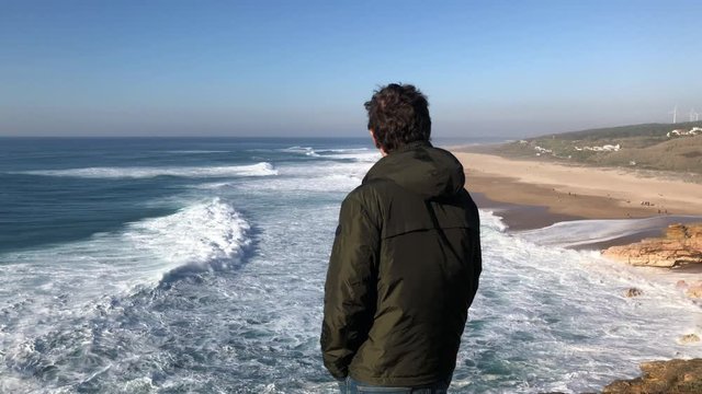 Young brunette man standing on steep cliff of Nazare coastline, enjoying a view of amazing crushing waves with white foam, drops of seawater; windy sunny evening; self-guided travel concept, Portugal