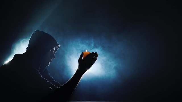 The silhouette of a man in a hood, holding a burning candle. Amid the swirling fog