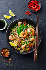Stir fry noodles with vegetables and shrimps in black bowl. Dark slate background. Top view.
