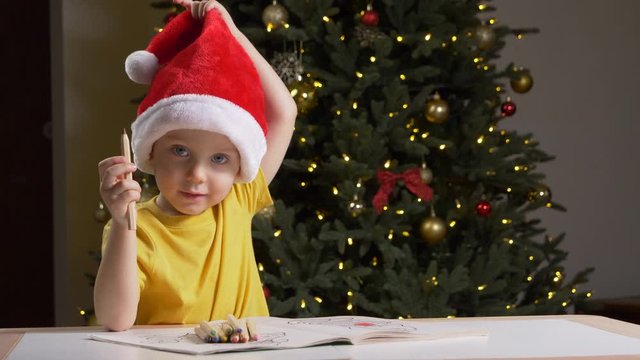  Cute child in Santa Claus hat is drawing with pencils in coloring book. Kids art, holidays activities, Christmas concept