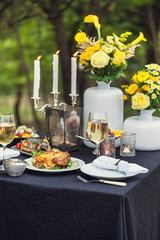 Baked full chicken with vegetables in a plate on a table next to glass of wine and candles. Outdoor, green park