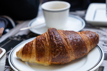 A well-done croissant on white dish in close up view. Blur cup on a saucer background.