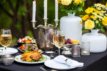 Baked full chicken with vegetables in a plate on a table next to glass of wine and candles. Outdoor, green park