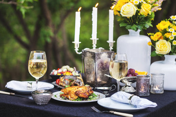 Baked full chicken with vegetables in a plate on a table next to glass of wine and candles. Outdoor, green park