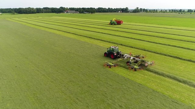 Aerial view of tractor creating windrows of grass for silage used for cow during winter in background showing combine harvester collecting the food and farm buildings further back 4k high resolution