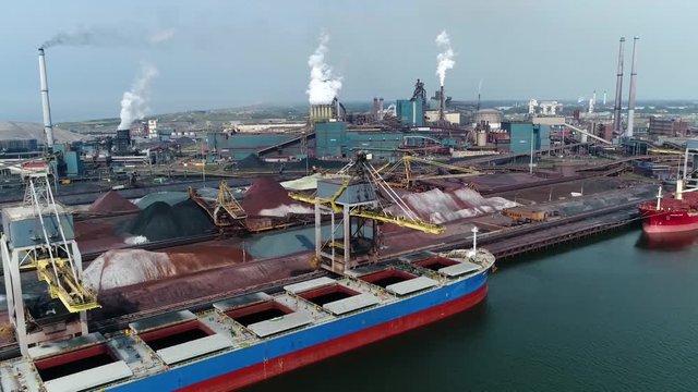 Aerial view of cargo ship unloading ore at steelmaking factory crane moving near vessel first showing the sluice of IJmuiden then the plant and the raw commodities already unloaded 4k high resolution