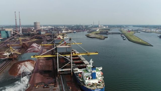 Aerial view of cargo ship unloading ore at steelmaking factory crane moving near vessel first showing the sluice of IJmuiden then the plant and the raw commodities already unloaded 4k high resolution
