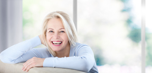 Active beautiful middle-aged woman smiling friendly and looking in camera near window at home....