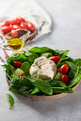 Italian cheese burrata with a salad of fresh spinach, cucumbers and cherry tomatoes with olive oil in a ceramic plate on a light background.