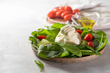 Italian cheese burrata with a salad of fresh spinach, cucumbers and cherry tomatoes with olive oil in a ceramic plate on a light background.