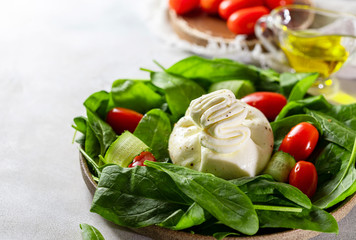 Italian cheese burrata with a salad of fresh spinach, cucumbers and cherry tomatoes with olive oil in a ceramic plate on a light background.