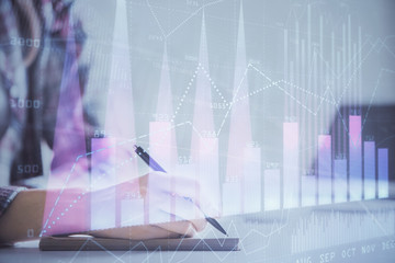 A woman hands writing information about stock market in notepad. Forex chart holograms in front....