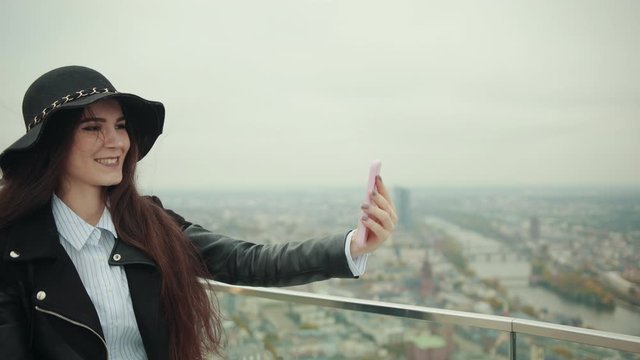Beautiful woman blogger in woman's hat takes video on the roof of a high building skyscraper a city metropolis and smiles at camera lens. Makes selfies