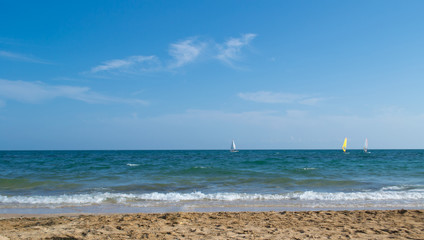 Sandy shore and blue-green sea against the blue sky. In the distance are sailboats.