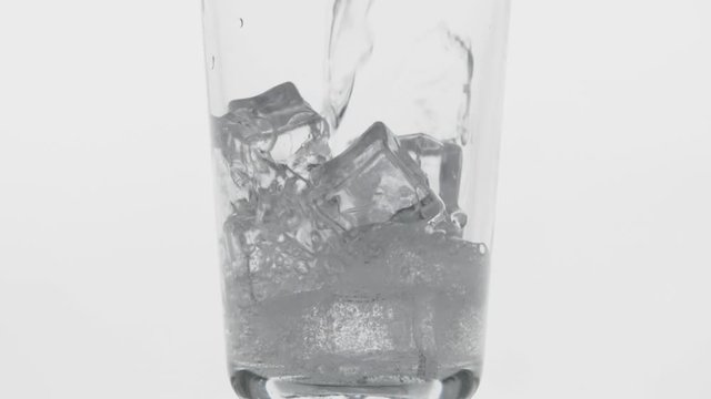 Close-up pouring transparent fizzy drink. Glass full of cold sparkling water with lce cubes on clean white background, moving bubbles in soda slow motion