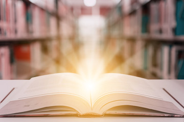 Rays of light coming from an open book or textbook on wooden table with blurred bookshelf in the library. Success education learning or back to school concept background