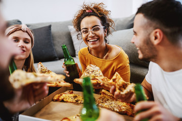 Group of friends sitting on the floor in living room, drinking beer and eating pizza. House party.