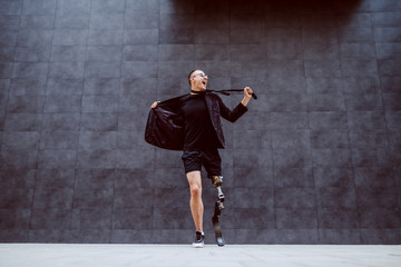 Full length of handsome Caucasian businessman with artificial leg getting tired of work and ripping his suit while standing outdoors in front of gray wall. It's time for sport.