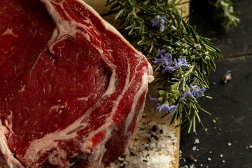 A raw beef chop on a chopping board with herbs and seasoning on a dark background