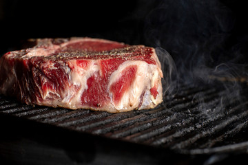 A beef chop cooking on a barbeque with smoke, on a dark background