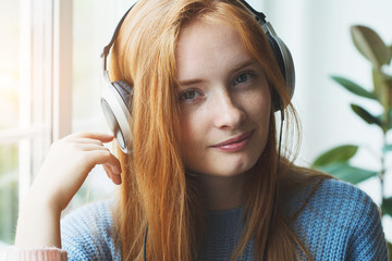 Fototapeta premium pretty smiling young red haired girl with headphones listening to music at home, relaxing, enjoying songs