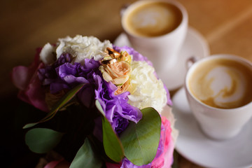 Close up of engagement rings, rose flower bouquet and coffee on table. 