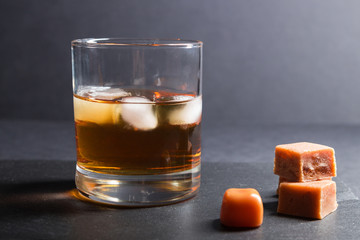 Glass of whiskey with ice and caramel candies on a black stone slate board on black background. Side view, selective focus.