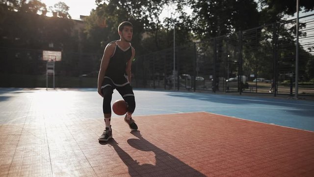 Close View of Caucasian Sporty Guy Trains to Play Basketball, Handling Ball, Bouncing Between his Legs and Throwing It Into Basket at Basketball Street Court . Healthy Lifestyle and Sport Concept.
