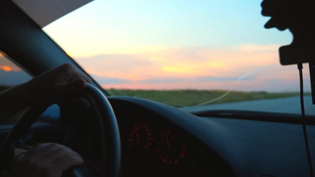 Unrecognizable man holding his hands on steering wheel of car and riding through countryside. Guy enjoying travel journey at evening time. Scenic view on road from inside auto. Slow motion Close up