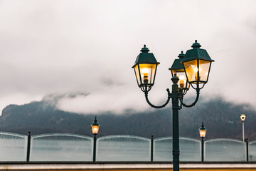 outdoor lantern yellow illumination waterfront walking district in foggy cloudy dramatic weather moody landscape background lake surface and mountains scenic view, empty copy space for your text