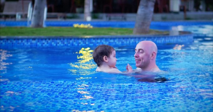 Father Holds the Baby in a Tight Embrace, They Splash in the Water, Dady Teaches the Little Son to Swim in the Pool. Cute Dad and Little Son are Smiling Happily.