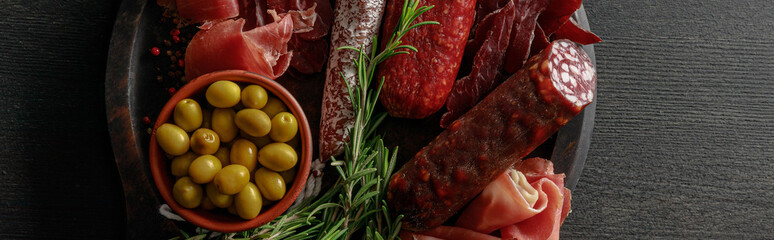 top view of delicious meat platter served with olives and herbs on wooden black table, panoramic shot