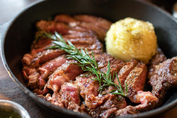 Cast iron plate with sliced grilled rib-eye cooking a medium-rare steak with mashed potato and sauce in saucer over table.