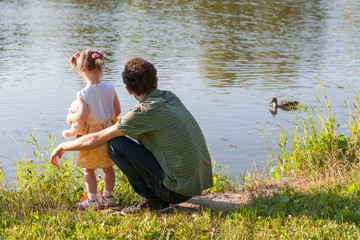 Father and daughter looking at lake. Girl is holding a Teddy bear. A wild duck swims through the water. Back view.