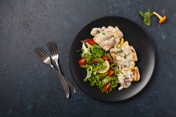 Fried cod served with mushroom sauce and salads. Portion on a black plate. Dark background.