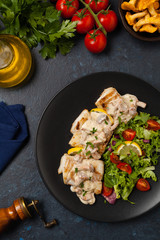 Fried cod served with mushroom sauce and salads. Portion on a black plate. Dark background.