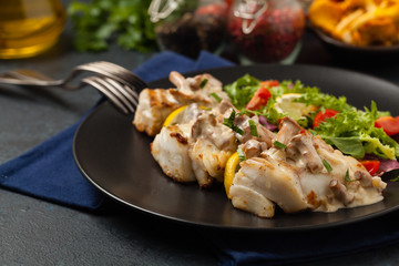 Fried cod served with mushroom sauce and salads. Portion on a black plate. Dark background.