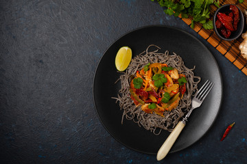 Wheat noodles with black sesame, fried in a wok with chicken and vegetables. Top view. Served on a black plate.